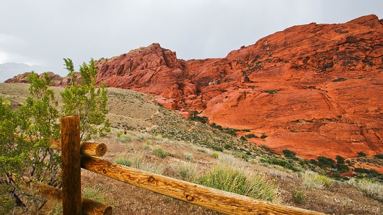 This picture features a striking landscape dominated by a vibrant red rock formation. The rock has a rugged texture and appears to be quite massive, stretching across the frame with various peaks and valleys. In front of the rock formation, there's a rustic wooden fence running horizontally, possibly marking a trail or property boundary. The foreground showcases a variety of desert shrubbery and grasses, hinting at a dry, arid climate. Overhead, the sky seems overcast with shades of gray, suggesting that it might be a cloudy or stormy day. Despite the dreary sky, the red rock's color remains vivid and is the focal point of the scene.