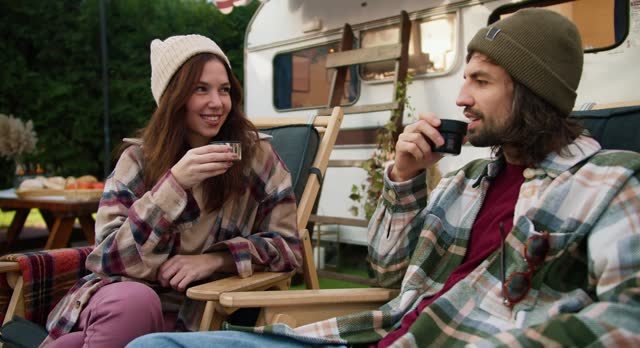 A happy brunette girl in a white cap and a checkered shirt drinks tea with her brunette boyfriend in a green checkered shirt sitting on special chairs during a picnic near her trailer in the forest in the summer
