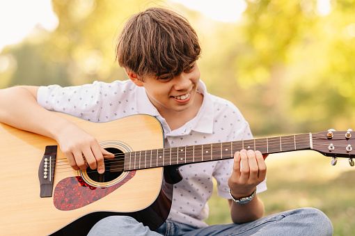 Cheerful young teen boy is happy he can play guitar while sitting in a park in summer.