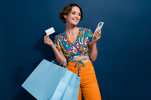 Photo of lovely young lady shopping bags gadget credit card dressed stylish flared pants garment isolated on dark blue color background.