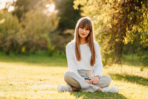 Full body shot of a blonde young woman with bangs sitting on grass relaxed in park on a sunny day.