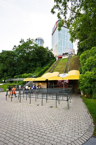 This is a vertical image featuring an outdoor scene with a funicular railway. In the foreground, there's a fenced area with cobblestone paving leading to an entrance with a covered ticket booth, surrounded by yellow umbrellas shading some tables. Beyond the ticket booth, a red funicular car is visible, ascending a steep, green hillside with tracks. In the background, on top of the hill, a tall glass skyscraper with red and white features dominates the skyline, with a shorter, cylindrical building to the left. The sky is overcast, suggesting a cloudy day. The area is surrounded by lush green trees, giving it a feeling of a park within a city.