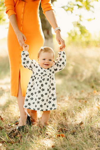 Wide smiling baby girl makes her first steps with the help of her mother outdoors.