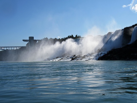 This image depicts a breathtaking view of a waterfall, most likely a section of the famous Niagara Falls based on the appearance and the observatory deck structure seen on the left. The powerful cascade of water plummets down, generating a mist that diffuses into the air, beautifully illuminated by the sunlight. The water's rush is so forceful that it leads to foam and turbulence where it meets the river below, which appears calm in the foreground.  The sky is mostly clear with hints of white clouds, giving the scene a vibrant and lively feel. On the left side of the falls, an observation platform with several levels allows visitors to experience the majestic view from a closer vantage point. A bridge can be seen in the distance connecting two land masses, partially obscured by the spray's mist. The combination of natural power and serene water with man-made structures suggests a harmony between nature and human engineering, inviting viewers to appreciate both the raw force of the falls and the opportunity to witness it up close through the provided facilities.