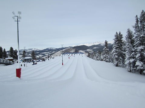 The picture shows a snowy landscape, likely a ski resort, with multiple well-groomed ski tracks leading down a slope. There are snow-covered trees on either side of the tracks, suggesting a mountainous area. Several skiers are visible in the distance, some queuing near a ski lift. In the background, majestic mountains with peaks dusted with snow extend across the horizon, indicating this location could be at a high altitude. The sky is overcast with a blanket of clouds, yet it appears to be a bright day, likely due to the high reflectiveness of the snow.
