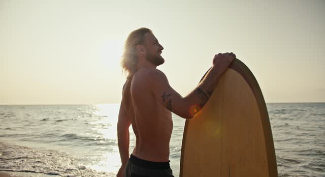 A blond man with a beard with a bare torso leans with one hand on his surfboard and with the other he covers his eyes from the sun and looks into the sea while standing on the shore in the morning at Sunrise