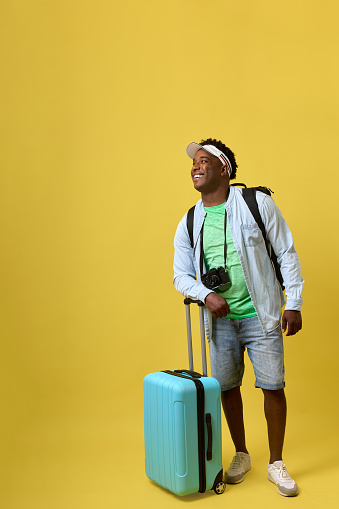 Happy African-American tourist, young man tourist in cap and denim shorts with suitcase and camera, posing on a yellow background. A smiling black guy with a suitcase is going on a summer trip