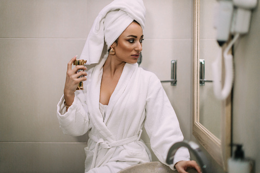 Beautiful young woman wearing a bathrobe and a towel applying perfume in a bathroom after showering