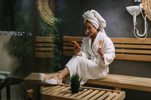 A beautiful young woman wearing a bathrobe and a head towel sitting in a hotel spa and using a smartphone