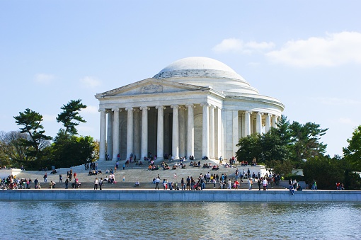 The picture shows the Jefferson Memorial, a neoclassical building in Washington D.C., United States. It is a bright day with a clear blue sky. The memorial has a low dome, surrounded by a circular colonnade of white columns. In front of the memorial, there is a body of water, which reflects the image of the structure. Several people are visible, scattered around the memorial's steps and promenade, suggesting a serene and tourist-friendly environment. Green trees flank either side of the memorial, adding a touch of nature to the architectural scene.