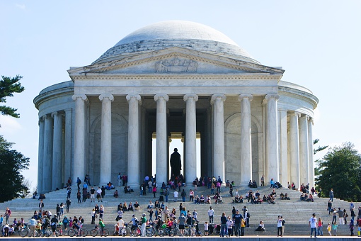 This image depicts the Jefferson Memorial, a famous landmark in Washington, D.C. The memorial is a neoclassical building with a large, white, circular dome supported by tall, slender columns. Capitals top each column, reflecting a Roman architectural style. The entrance is reached via a broad set of stairs, where many visitors are seen sitting and standing. Inside the portico, a silhouette of a statue is visible. The sky is clear with a few soft clouds, indicating a sunny day, which casts natural light on the memorial's white facade, highlighting its grandeur. The ground in front of the memorial appears to be a paved promenade with some visitors walking, and there's greenery to the sides. Overall, the scene conveys a sense of historical significance and serves as a popular gathering place for tourists and locals alike.