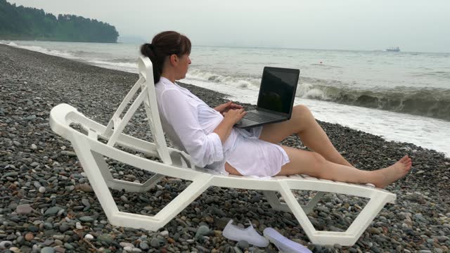 A woman lies on a sun lounger with a laptop on the seashore.