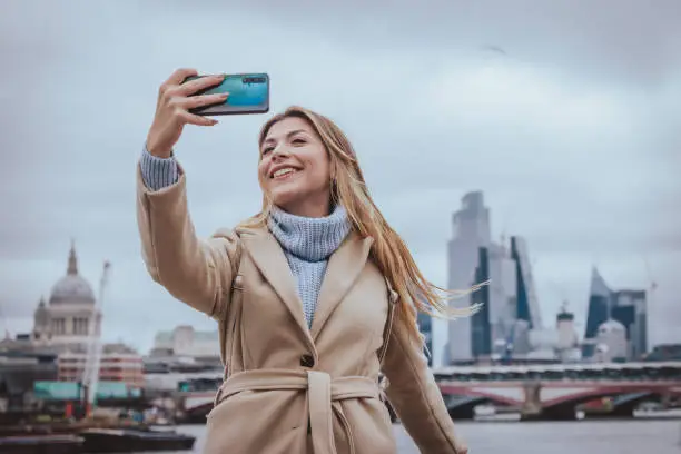 Candid portrait of a young cheerful blond girl in her 30s taking selfie with her smart mobile phone while smiling, as background of city of London's skyline, England, UK. Selective focus on the model with plenty of copy space at the background, which is defocused Saint Paul’s cathedral, Blackfriars Bridge and all modern business buildings at the heart of the capital. Photo created during cold season outdoors and the model is with warm casual clothes on a cloudy day  - creative stock photo
