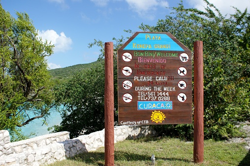 The picture shows a large wooden signboard at a beach entrance. The sign is primarily in a reddish-brown color with white text. It reads 