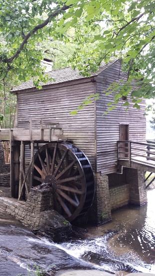 The picture shows an old-fashioned wooden mill nestled beside a creek or small river. The focal point of the image is a large, dark wooden waterwheel attached to the side of the mill, partially in the water, suggesting that it is powered by the flow of the stream. The building has a rustic charm, with weathered wooden siding and a shingled roof. A small wooden bridge or walkway connects the mill to the creek bank, allowing access to the structure. Trees with green foliage gently overhang the scene, providing a sense of tranquility and natural beauty. The water of the creek appears to be moving, with some ripples visible, and the setting appears to be peaceful and secluded.