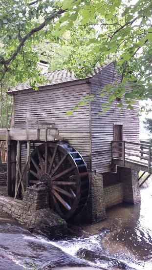 This is a photo of a traditional watermill, which is a structure typically built near a water source such as a river or stream. The watermill depicted here is made of wood, with what appears to be weathered and rustic wooden siding, indicative of its age and the natural elements it has endured. It has a somewhat sloped roof, possibly covered in shingles, which is common in mill construction.  The most prominent feature of this watermill is its large waterwheel, which is also made of wood. The wheel is positioned vertically and appears to be in good condition, with all its slats intact. It is positioned partially inside the water, which would traditionally turn the wheel to power the internal mechanisms of the mill used for grinding grain or other materials.  The watermill is positioned atop a small stone foundation that raises it above the flowing water. You can see the water gently flowing past the bottom of the mill, indicating that the water level is not too high, perhaps during a drier season or time of day.  There are lush green leaves from the surrounding foliage overhanging the scene, which provides a sense of tranquility and indicates that the watermill is located in a green, wooded area.  Overall, the image evokes a sense of historical technology, rural life, and peaceful natural beauty.