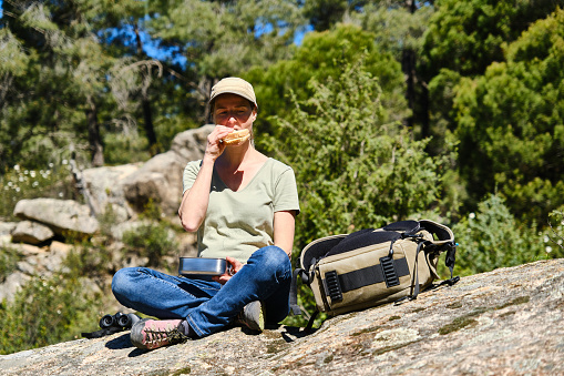Young female traveler in jeans and cap sitting on stone with backpack while eating sandwich during trip against blurred trees of green forest