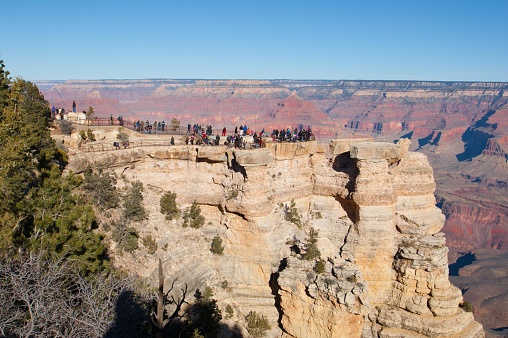 This is a picture of a group of people at the Grand Canyon National Park, standing on an observation deck that juts out from a cliff. The deck offers a panoramic view of the canyon with its layered bands of red rock that reveal millions of years of geological history. The sky is clear and blue, emphasizing the vastness and depth of the canyon. The terrain is rugged with sparse vegetation, and the natural light is bright, suggesting the photo was taken during the day. The crowd appears to be enjoying the view and the experience of visiting one of the natural wonders of the world.
