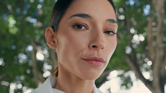 Portrait of calm elegant woman standing in front summer greenery alone. Attractive young businesswoman looking camera seriously outdoors. Confident chic lady brunette with stylish hairstyle close up.