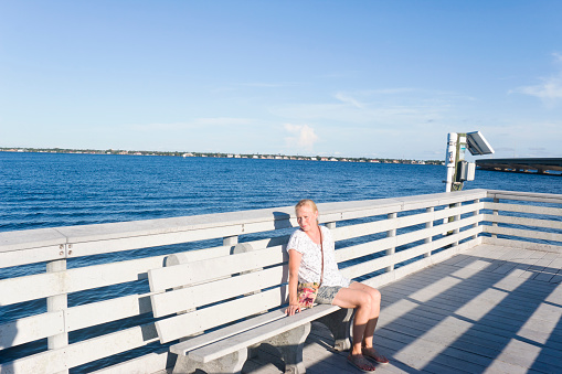 Blonde woman on a bench at a fishing pier at the Four Mile Cove an ecological preserve at the Caloosahtchee River in Cape Coral, Florida, USA. The Caloosahtchee River flows in the Gulf of Mexico.