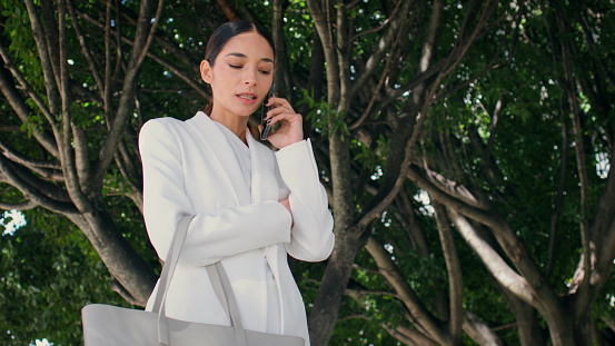 Successful business woman calling telephone standing under beautiful city trees in elegant white suit. Confident chic businesswoman discussing work issues by phone. Beautiful girl talking on cellphone