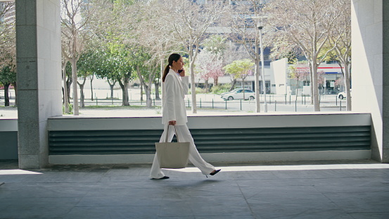 Chic business lady calling hurrying in office wearing trendy white suit. Glamorous young businesswoman discussing work using smartphone outdoors. Elegant woman talking telephone going in building.
