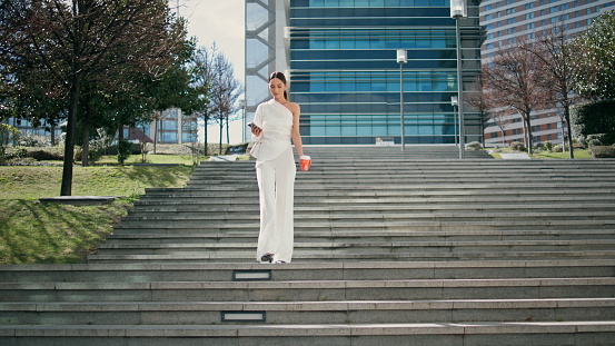 Elegant woman going downstairs looking on smartphone screen sunny day. Chic young businesswoman messaging on telephone walking down staircase with coffee cup. Luxurious lady using phone outdoors.