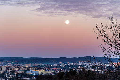 Brno, Czech Republic - January 2, 2024: Shining full moon in the evening blue sky above the skyline of city buildings.