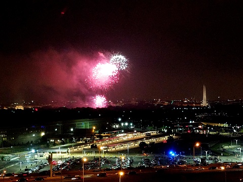 This is a nighttime image taken from an elevated perspective, showing a cityscape with fireworks exploding in the sky. The fireworks are mostly in shades of pink and white. In the background, on the right side of the image, the iconic Washington Monument is visible, illuminated and standing out against the dark sky. In the foreground, there's a well-lit parking lot full of cars, and various buildings with interior lights on. The city glows with both the celebratory fireworks and the bustling activity of the urban environment.