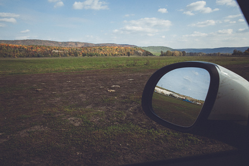 An image of a car side mirror with reflection of a field with sparsely growing grass and shrubs, and distant mountains, taken on a road trip on a sunny day with a blue sky and white clouds