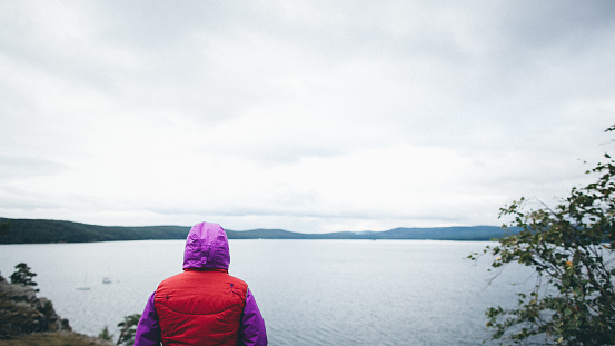 A woman standing on the rocky shore of a lake, gazing out at the water on a cloudy day, wearing a purple-red jacket, appearing contemplative, with mountains in the background and cloudy sky.