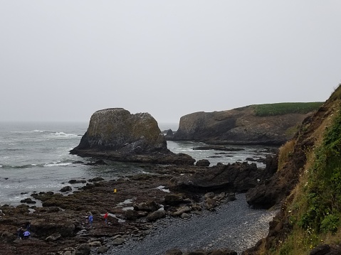 This image reveals a coastal scene where the vast, churning sea meets a rugged shoreline. A series of prominent, rocky outcrops rise majestically from the water, showcasing the force of erosion sculpting the landscape. The sky is overcast with a blanket of grey clouds, suggesting a cool and misty atmosphere. The rocky shore is dotted with tidal pools and scattered rocks. On the left, vibrant greenery clings to the cliff, possibly indicating some resilient coastal flora. Down below, a few people can be seen exploring the area, appearing quite small in comparison to the grandeur of the natural surroundings. The overall mood is one of solemnity and the raw beauty of nature.