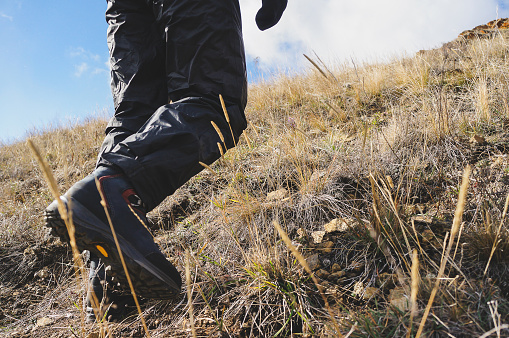 A lone adventurer in black pants and sturdy brown boots conquers a rugged slope with dry grass, against a backdrop of a majestic mountain range shrouded in ominous dark clouds.
