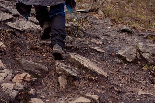 A person in black hiking boots and blue pants confidently hikes on a rocky trail in the woods, surrounded by trees and shrubs. Ground covered with rocks, dirt, and leaves. Low angle shot.