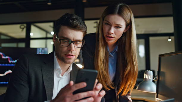 istock Two business people working dark finance office. Focused man using smartphone 1998548540