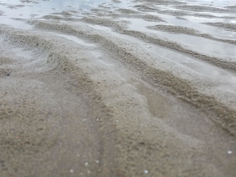 This is an image of a sandy beach with a low tide, which has left behind shallow pools of water in between ridges of sand. The sand has a rippled texture, likely created by the movement of the water. The sand's color is a light brown, and the water appears to be clear. The perspective is close to the ground, giving a sense of the textures and patterns in the sand. The atmosphere looks overcast as the lighting is soft and diffused, without harsh shadows, suggesting an overcast or cloudy sky.