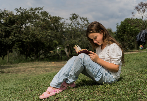 Portrait of a beautiful girl sitting outdoors on the grass while concentrating on reading a book.
