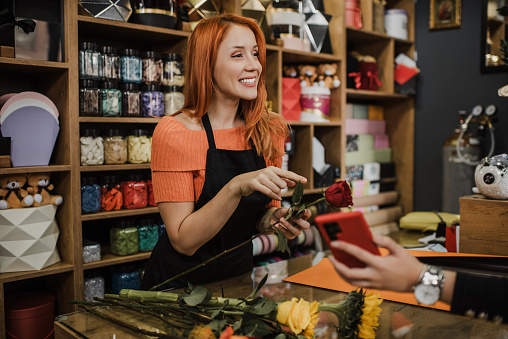 Beautiful redhead female seller working in modern flower shop. She is happy and helps her customer to make the right choice of flowers. Small business concept.