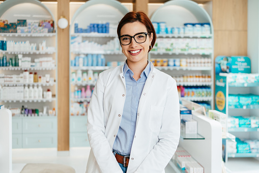 Beautiful pharmacist working and standing in a drug store and doing a stock take. Portrait of a positive healthcare worker or a chemist at his work. She is standing and looking at camera.