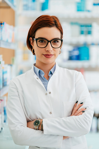 Beautiful pharmacist working and standing in a drug store and doing a stock take. Portrait of a positive healthcare worker or a chemist at his work. She is standing and looking at camera.