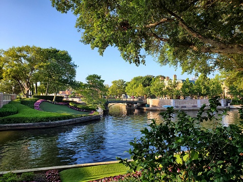 The image depicts a tranquil urban park scene. A clear blue sky hovers over a serene body of water that reflects the surrounding foliage and structures. A stone bridge with balustrades spans the water, connecting two lush green banks. The banks are adorned with manicured grass, flowering plants in a linear arrangement, and mature trees providing dappled shade. To the left, a gentle slope leads down to the water's edge. The environment suggests a well-maintained, peaceful public space, likely within a city due to the buildings visible in the distant background. The overall composition of the image conveys a sense of calm and natural beauty within an urban setting.