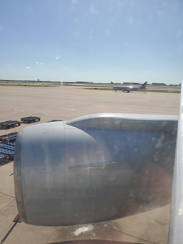This image shows a view from an airplane window. Looking out onto the tarmac, we can see the large, grey engine of the airplane, part of the wing, and another aircraft in the distance, likely taxiing on the runway. The skies are clear with just a few clouds, suggesting good weather conditions for flying. There is also baggage handling equipment visible towards the left, indicating the proximity to an airport terminal or baggage loading area.
