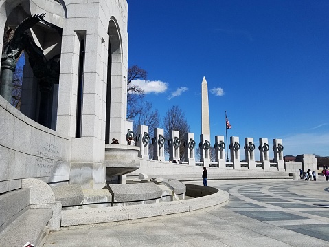 The picture shows the World War II Memorial located in Washington D.C., USA. It's a sunny day with a clear blue sky. The memorial consists of a circular arrangement of pillars, each representing a U.S. state or territory at the time of World War II. In the center, there are two large arches, and in the foreground, there's a broad stone pavement with a few steps leading up to the memorial. The Washington Monument, a tall and narrow obelisk, is visible in the background, centered behind the memorial. Several American flags are displayed around the circumference of the memorial, gently waving in what appears to be a light breeze. There are a few visitors scattered around the scene, some walking and others sitting by the pillars. The trees around the memorial are mostly bare, suggesting it might be either late fall or early spring.