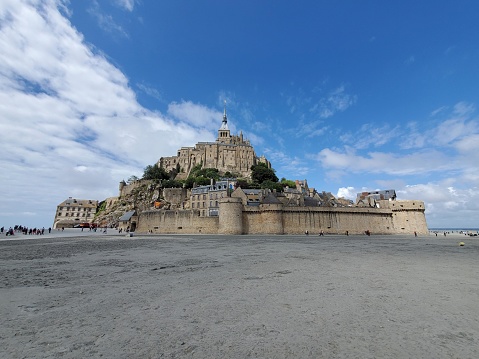 This picture shows Mont Saint-Michel, an iconic island commune located in Normandy, France. The island features a medieval village at its base and a historic abbey at its peak. The architecture is of a Gothic style, with the abbey's spire reaching toward the sky. The image captures the structure in clear weather, under a blue sky with scattered clouds, emphasizing its grandeur. Around the base, you can see the fortifications including walls and towers. People appear to be walking on the expansive flatland around the island, which suggests that the tide is low, making the sandy bottom accessible to visitors. Mont Saint-Michel is renowned for these dramatic tides, which can vary greatly and quickly surround the island with water. Overall, this is a breathtaking and imposing view of a significant historical landmark.