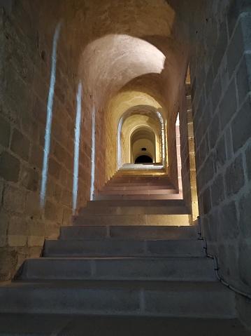 The picture depicts a series of stone steps leading up through a narrow passageway. The walls on either side are made of large, coarse stones that add a rustic feel to the environment. The pathway is dimly lit, which creates an ambiance of tranquility and mystery. The walls have some openings or indentations casting light, suggesting there might be some other chambers or sources of light adjacent to the stairwell. The vanishing point is at the top of the stairs, drawing the viewer's attention upward, making one curious about what lies at the end of the staircase.