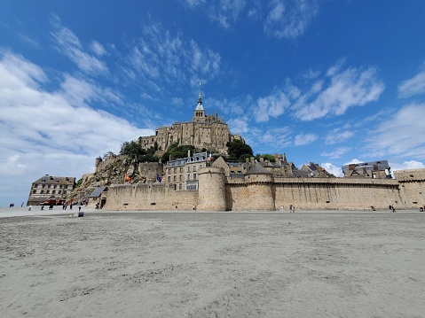 This picture shows the magnificent Mont Saint-Michel, a historic fortified island commune located in Normandy, France. The image captures a clear, bright day with a few clouds scattered across the bright blue sky. On the island, the structures rise progressively with the lower part consisting of sturdy stone walls and traditional buildings, leading up to the prominent abbey and monastery at the top, crowned with a spire and cross.  The expansive flat sand surrounding Mont Saint-Michel suggests that the tide is out, revealing the tidal flat area. A few people can be seen in the distance walking on these flats, emphasizing the vastness of the area and the imposing stature of the Mont. The architectural details of the buildings imply a rich history and a combination of military, religious, and residential functions over the centuries.
