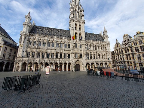 The picture showcases a stunning view of the Grand Place, also known as Grote Markt, in Brussels, Belgium. This historic square is surrounded by opulent guildhalls and other edifices with intricate facades featuring ornate stonework and decorative elements. The dominant building in the view is the Brussels City Hall, distinguished by its Gothic architecture and soaring bell tower topped with an intricate spire. The square is paved with stones and appears almost empty, giving it a tranquil atmosphere. The overall scene is under a clear sky with few clouds, suggesting a pleasant day, ideal for visiting and appreciating the architectural beauty of this UNESCO World Heritage site.