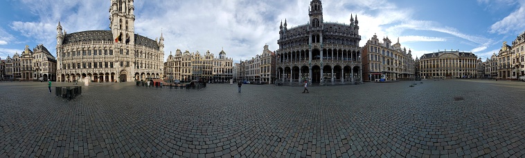 This is a panoramic photo of the Grand Place (Grote Markt) in Brussels, Belgium, a UNESCO World Heritage Site. The picture shows the expansive cobblestone square flanked by ornate, historic buildings with opulent architecture that showcases the city's rich history and legacy. In the background, the sky is mostly clear with a few scattered clouds, and it's during daytime as indicated by the natural lighting and shadows on the ground. Visible is the towering Town Hall with its gothic style and spire, which dominates the skyline. Surrounding the square are guildhalls and other stately structures with intricate facades, embellished with statues, gilding, and stonework. Despite the grandeur surrounding the square, there are few people, suggesting it might be a quiet moment for this usually bustling center. The absence of modern elements keeps the focus on the historic and architectural beauty of the setting.
