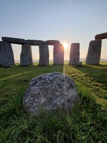 This is an image of Stonehenge, an iconic prehistoric monument located in Wiltshire, England. The picture captures the site at what appears to be either sunrise or sunset, as indicated by the low position of the sun in the sky, creating a beautiful starburst effect as it peers through the gaps between the standing stones.  The stones are seen in silhouette and are large, with some arranged in a circle and others positioned on top of these vertical slabs, forming lintels, creating the well-known trilithon structure. One large, flat stone is visible in the foreground, possibly one of the fallen sarsen stones or part of the altar stone, and the well-maintained grass that surrounds the stones suggests it's early morning dew or well-irrigated grass. The sky has a gradient of colors, fading from the warm tones near the sun to a clear light blue as it stretches upwards. The image captures the mystery, historical significance, and natural beauty of this ancient site.