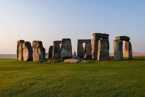 The picture shows Stonehenge, a prehistoric monument in Wiltshire, England. It consists of a circular setting of large standing stones set within earthworks. In this image, the monument is seen during daylight with a clear sky. The grass around the stones is a lush green, indicating the photo was likely taken in spring or summer. Shadows cast by the stones suggest the sun is low in the sky, possibly during early morning or late afternoon. Stonehenge is one of the most famous landmarks in the United Kingdom and a UNESCO World Heritage Site.